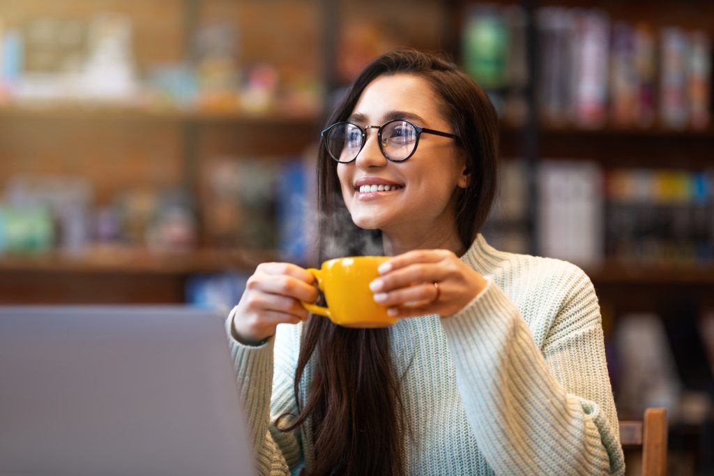 Benefits of remote work. Dreamy lady enjoying coffee during work on laptop in city cafe, smiling and